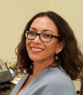 Bobbie Rosa, a young Native American woman, with shoulder length brown hair, rectangle glasses, and an eyebrow ring, smiles for a photo over her shoulder.