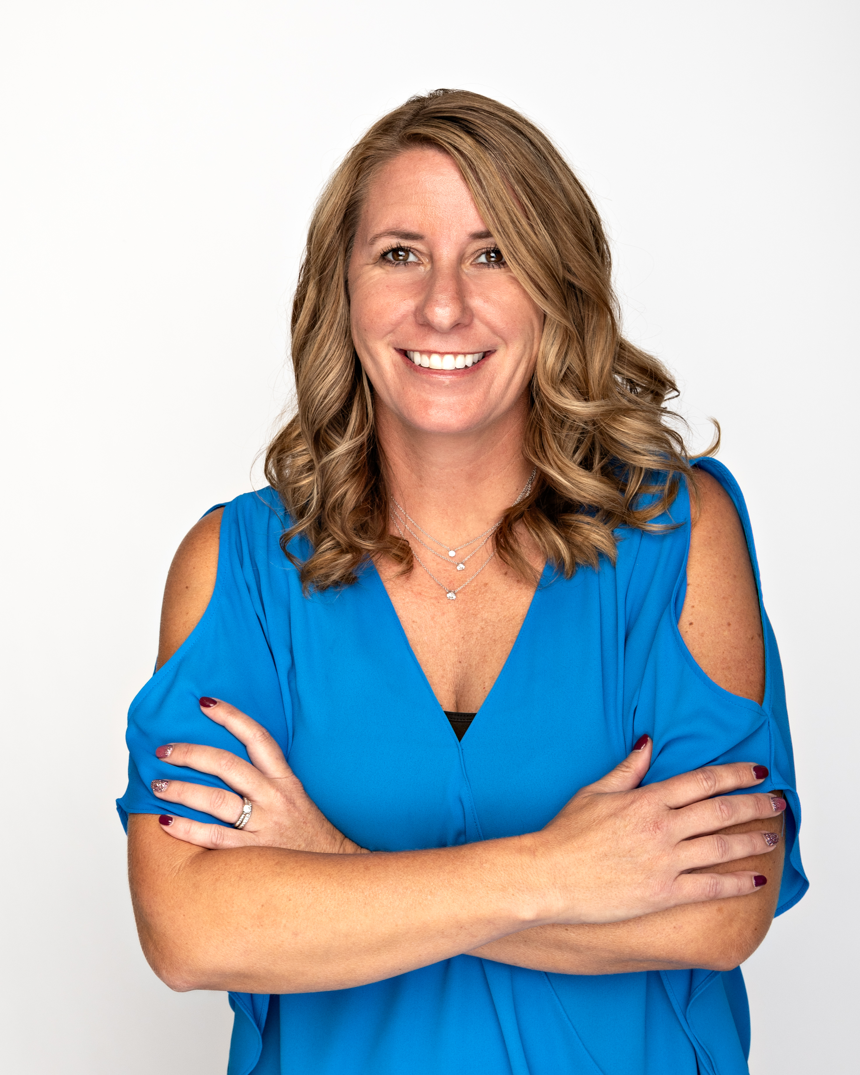 Katie, a young white woman with shoulder length curly light brown hair, wears a blue cold shoulder top, and smiles in a headshot.