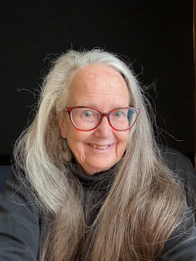 Amy, a light skinned woman with long gray hair and red glasses smiles against a dark background.