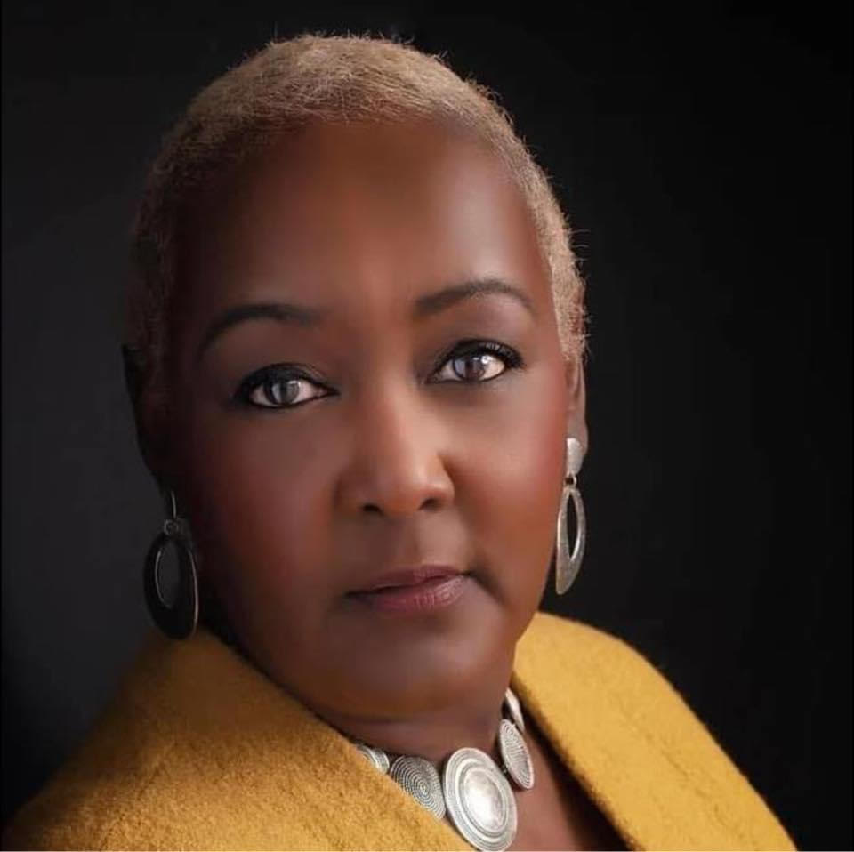 Yolanda, a middle aged Black woman with short brown hair, wearing striking sterling earrings, necklace, and a yellow wool coat, poses for a headshot.